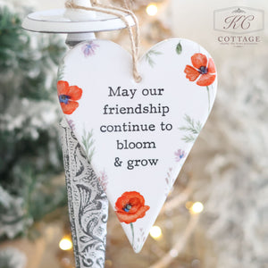 The Ceramic Poppy Hanging Heart with Friendship Quote, perfect as a gift for a friend, is elegantly displayed on a decorative stand. It features red flowers and the heartfelt message: "May our friendship continue to bloom & grow." The backdrop of blurred lights and foliage adds to the cozy atmosphere.