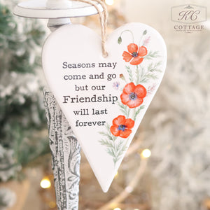 The Ceramic Poppy Hanging Heart with Friendship Quote features a heart-shaped design with red flowers and green leaves, offering the message "Seasons may come and go but our friendship will last forever." With a blurred Christmas tree backdrop, it makes an ideal gift for a friend.