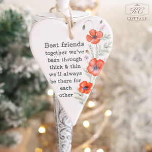 The Ceramic Poppy Hanging Heart with Friendship Quote boasts floral designs and reads, "Best friends together we've been through thick & thin we'll always be there for each other." It makes an ideal gift for a friend, featuring a backdrop of softly blurred lights and greenery.