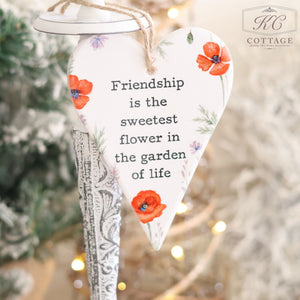 A Ceramic Poppy Hanging Heart with Friendship Quote, featuring red poppies and green leaves, beautifully displays the message, "Friendship is the sweetest flower in the garden of life." This thoughtful gift for a friend hangs gracefully against a backdrop of softly blurred lights and greenery.