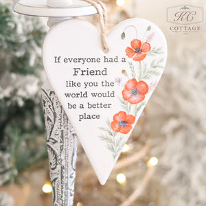 The Ceramic Poppy Hanging Heart with Friendship Quote showcases red flowers and green leaves, accompanied by the message, "If everyone had a friend like you, the world would be a better place." It is displayed against a soft-focus backdrop featuring a tree and the KC Cottage logo in one corner. This piece is perfect as a gift for a friend.