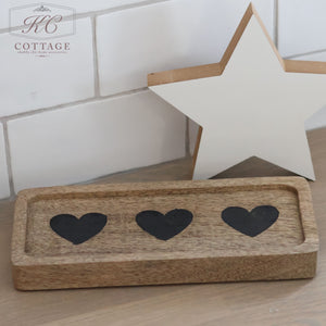 A Rectangle Rustic Wooden Tray, featuring three black hearts, exudes rustic charm as it sits on a wooden counter. Behind the tray, a decorative white star with a wooden border leans elegantly against the subway-tiled wall, completing an enchanting Hearts & Stars design.