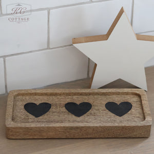 The Rectangle Rustic Wooden Trays - Hearts & Stars exudes rustic charm with its design featuring three black heart cutouts. It rests on a wooden surface, while a white star-shaped decoration leans against the white tiled wall, highlighting the enchanting hearts and stars theme.