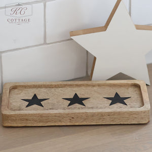 The Rectangle Rustic Wooden Tray, adorned with three black stars, exudes rustic charm as it rests on a wooden surface. Behind it, a white star-shaped ornament leans against a white tiled wall, perfectly embodying the Hearts & Stars design theme.