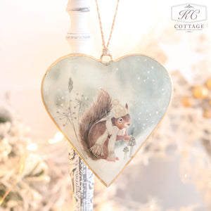 The Christmas Metal Woodland Animal Heart Hanger features a delightful design of a squirrel in winter clothes, sitting on a snow-covered ground alongside woodland mushrooms. Its light blue background decorated with snowflakes makes it an ideal addition for adding seasonal charm to your festive home decor.