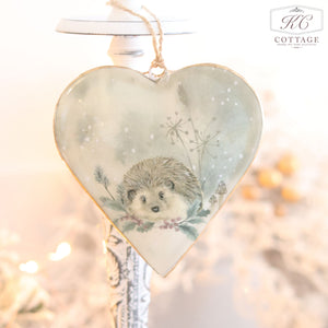 This heart-shaped ornament, known as the "Christmas Metal Woodland Animal Heart Hanger," features a charming hedgehog nestled among intricate foliage and snowflakes. It hangs gracefully from a string against a softly lit, blurred background, making it a perfect festive home decor piece.