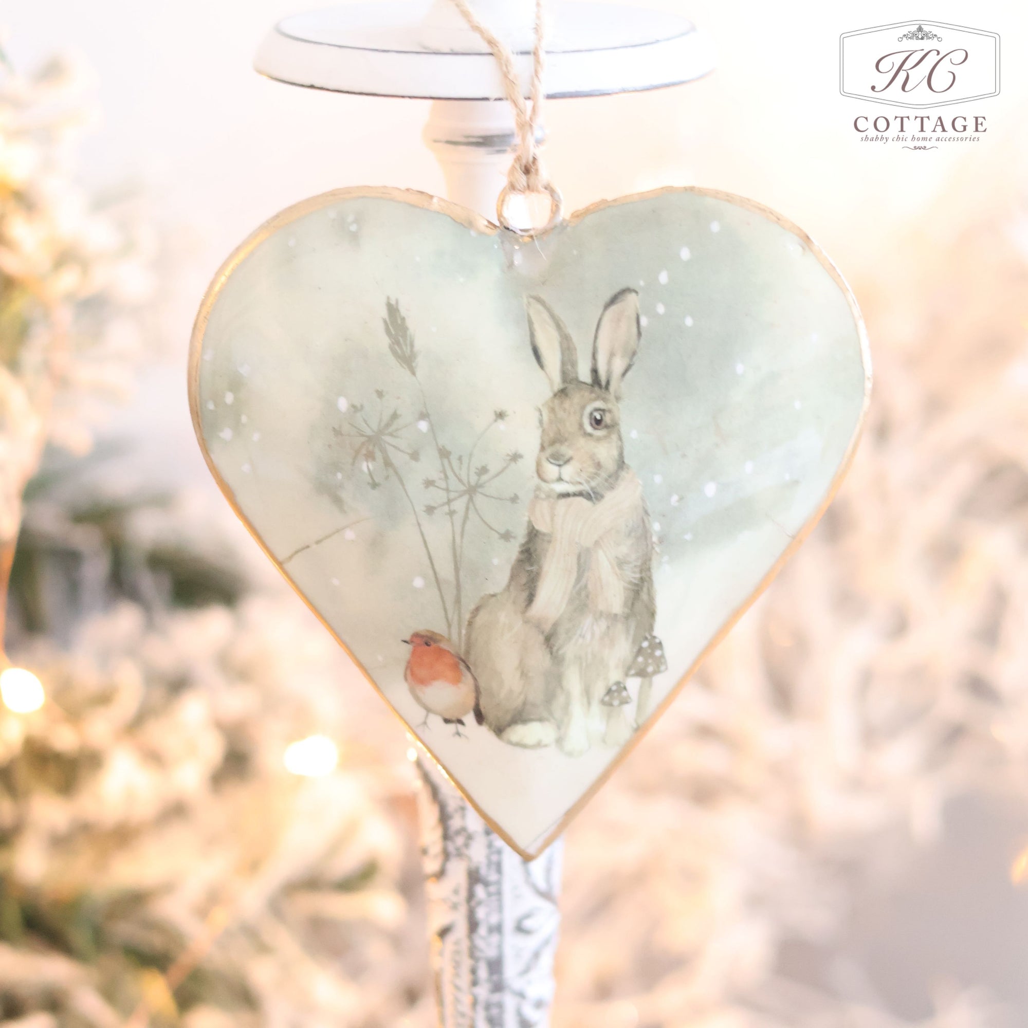 This Christmas Metal Woodland Animal Heart Hanger showcases an enchanting heart-shaped design with a delightful illustration of woodland animals, including a rabbit and a small bird, set against a snowy background. Hanging from rustic string, it adds festive charm to your home decor, complemented by the soft glow of a holiday tree in the background.