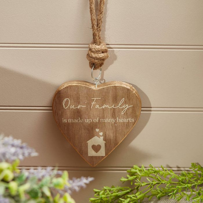 A Mango Wood Our Family Heart Hanger, engraved with "Our Family is made up of many hearts," hangs by a rope against a beige, paneled background. The ornament features a small house design and in the foreground, out-of-focus green and purple foliage adds a natural touch to the home decor.