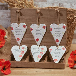 A wooden display holds six Ceramic Poppy Hanging Hearts with Friendship Quotes, making it an ideal gift for a friend. The Ceramic Poppy Hanging Hearts with Friendship Quotes are suspended by twine and arranged in two rows, with bright red poppies scattered around the base.