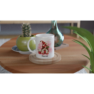 A white Personalised Christmas Gonk Mug sits on a coaster on a wooden table. The customisable mug features a design of a red-roofed house with plants and the word "Name" below the illustration, adding a festive touch. Also on the table are a potted cactus, a green vase, and a plant leaf in the foreground.