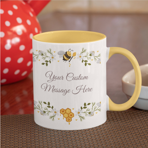 A white and yellow coffee mug on a table features a decorative bee and floral design. The Personalised Bee Mug Fully Custom Message has a customizable message area, surrounded by green leaves, yellow flowers, and honeycomb patterns. A red teapot with white polka dots adds charm to the home decor in the background.