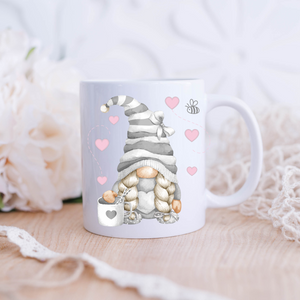This Grey Love Gonk with Hearts Mug showcases a cute gnome illustration with long braided hair and a striped hat. The gnome is holding a coffee cup adorned with a heart. Pink heart doodles and a small bee surround the gnome, set against a background of soft flowers and lace details—perfect for home decor.