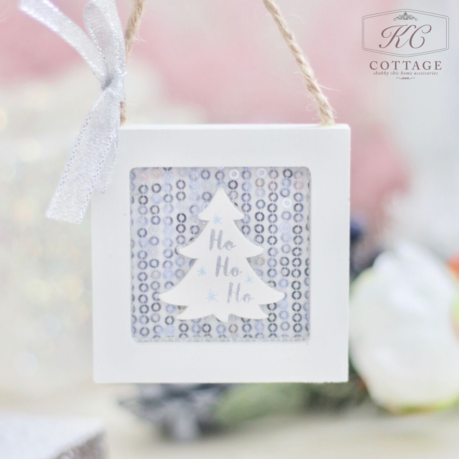 Introducing the Christmas Wooden Sequin Hanging Decoration: this elegant piece features a white Christmas tree silhouette with "Ho Ho Ho" beautifully incorporated within a square frame. It hangs from twine, set against a softly blurred background that highlights subtle festive colors, perfectly capturing the cheerful holiday spirit.