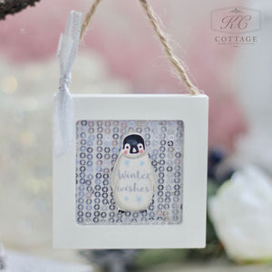 The Christmas Wooden Sequin Hanging Decoration features an adorable penguin illustration with the words "Winter Wishes," enclosed in a white square frame. This charming piece, perfect for Christmas, hangs with twine and ribbon against a softly blurred pink and white backdrop accented with light bokeh for extra festive charm.