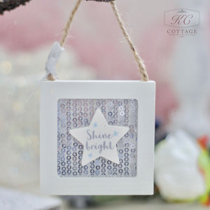 The Christmas Wooden Sequin Hanging Decoration, featuring a white star and the phrase "Shine bright" on a shimmering sequin background, brings an enchanting touch to your holiday decor. Its festive design is beautifully enhanced with a rustic twine hanger, set against a soft, blurred backdrop.