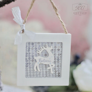 The Christmas Wooden Sequin Hanging Decoration showcases a reindeer silhouette with "Believe" inscribed, elegantly framed by a white border. This decorative piece is adorned with sequins and suspended from a rope embellished with a small silver bow, set against a softly blurred background that captures festive shapes and charm.