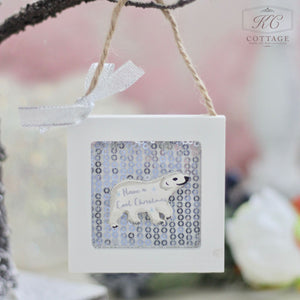 The Christmas Wooden Sequin Hanging Decoration features a small polar bear with the phrase "Have a Cool Christmas." It is set within a white square frame against a sequined backdrop, and it hangs from a tree branch by a string embellished with a white ribbon bow.