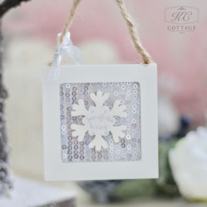 The Christmas Wooden Sequin Hanging Decoration is a square ornament showcasing a white snowflake on a glittering silver background. The phrase "snowflake kisses" graces the snowflake, complemented by festive shapes. It comes with a rope and ribbon for hanging, making it an ideal addition to add sequin-like sparkle to your holiday decor.