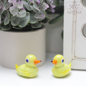 Two small glass duck ornaments with orange beaks and blue eyes are placed on a white surface. They are positioned near a potted plant with green and purple leaves and a white decorative frame, adding charm to your home decor.