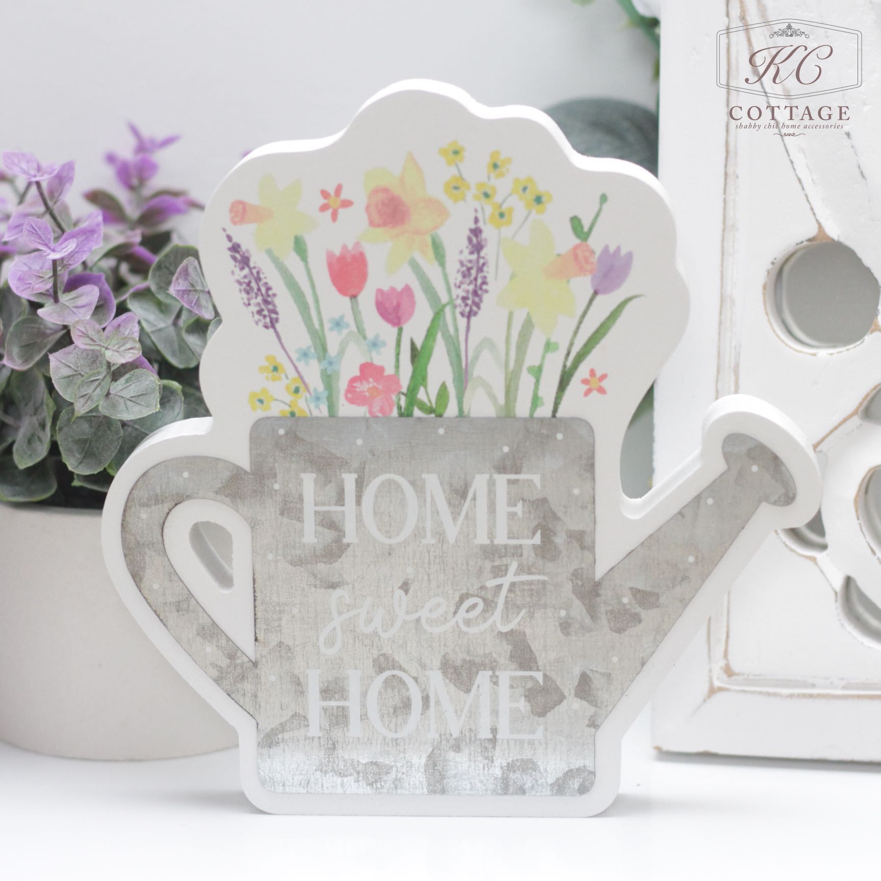 A Home Sweet Home Wooden Watering Can with floral designs at the top and the words "Home Sweet Home" written on it. Purple flowers and greenery are in the background, complemented by a rustic white decor piece on the right. Perfect for enhancing your home decor.