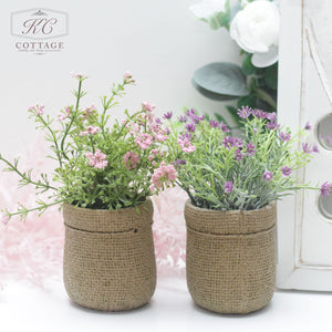 Two small Faux Spring Flowers in Hessian Pot sit alongside each other on a white surface. Behind them, a white decor item and a blurred background featuring white flowers are visible. The text “KC COTTAGE” appears in the top left corner, enhancing the cottage style charm.