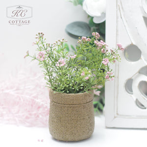 A small Faux Spring Flowers in Hessian Pot holds delicate faux spring flowers and green foliage. The background features a white, ornate decorative panel and soft-focus greenery. The image has a subtle, cottage style aesthetic, with the logo "KC Cottage" in the top left corner.