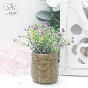 A Faux Spring Flowers in Hessian Pot sits on a white surface. The background features a decorative white cutout panel and soft, out-of-focus greenery. A logo in the top left corner reads "KC Cottage" with a delicate design, capturing the essence of cottage style.