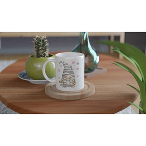 A Grey Love Gonk with Hearts Mug with an illustration of a gnome wearing a striped hat and holding a heart sits on a wooden coaster on a round wooden table. Nearby, there's a small cactus in a green pot and a tall, teal glass vase. Green leaves frame part of this charming home decor scene.