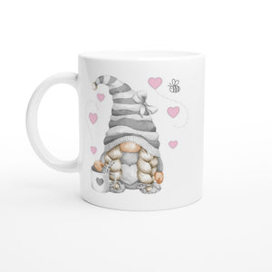 This charming Grey Love Gonk with Hearts Mug, perfect for home decor, showcases a cute gnome with long braids, a striped hat, and heart-patterned clothing. The gnome holds a heart-decorated mug while pink hearts and a buzzing bee add a whimsical touch.