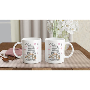 Two Grey Love Gonk with Hearts Mugs featuring gnome illustrations with gray striped hats, holding orange hearts. Pink hearts and bees are also part of the design, making these mugs charming pieces of home decor. They rest on a checkered tablecloth, with white flowers and a pink notebook in the background.
